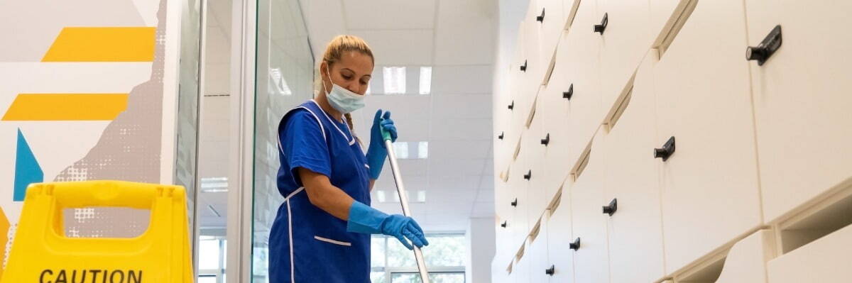 A cleaner with a mask on her face cleans the floor with the mop.Caution wet floor sign close up